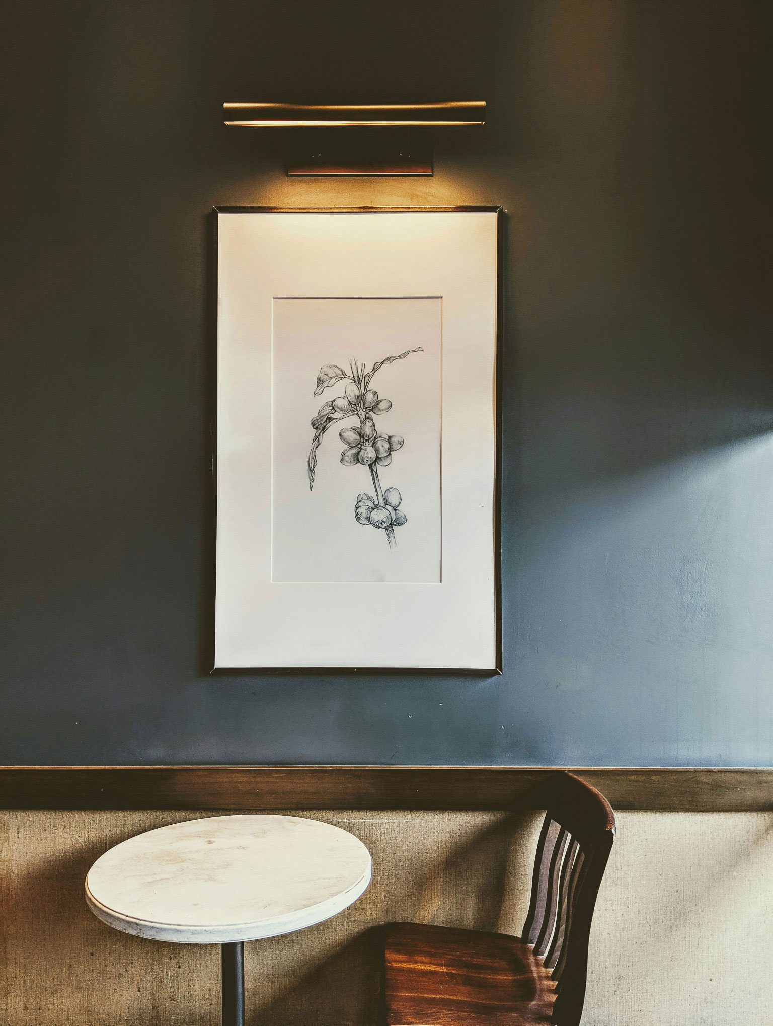 darshan-patel-LWNKjKmxRrE-unsplash A solitary round table with a wooden chair is positioned beneath a framed botanical sketch illuminated by an overhead light on a dark-toned wall.