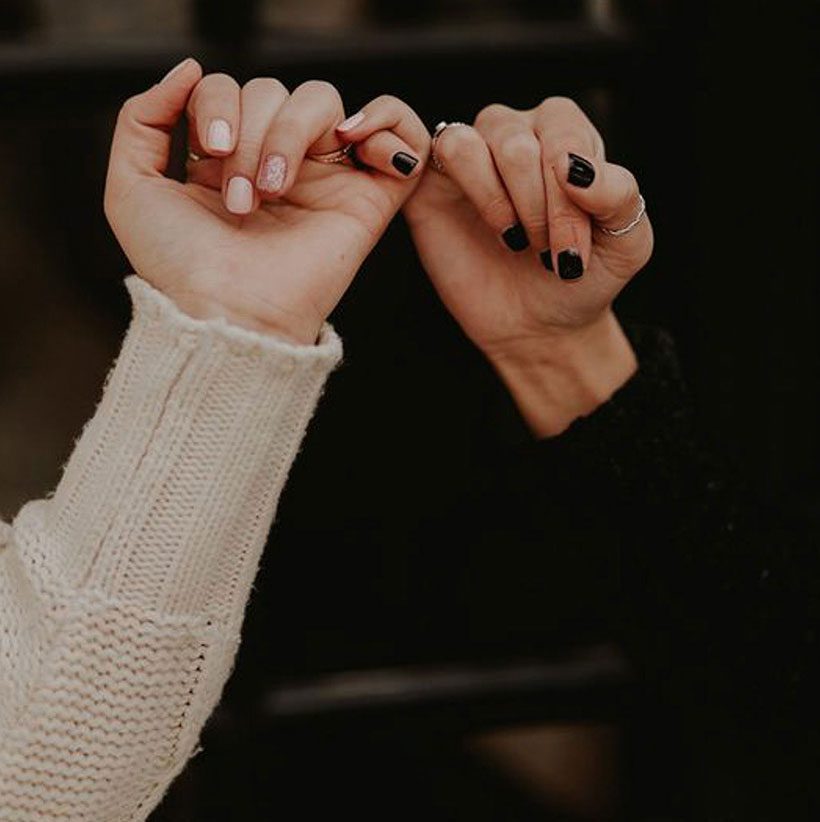 home-bottom Two hands with different nail polish colors make pinky promises against a dark background. One hand has light-colored nails and a cream sweater; the other has black nail polish and a black sleeve.