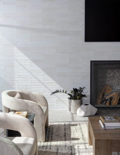 A modern living room with light-colored armchairs, a wooden coffee table, a fireplace with a plant beside it, and a wall-mounted TV above. Sunlight casts shadows on the white tiled wall.