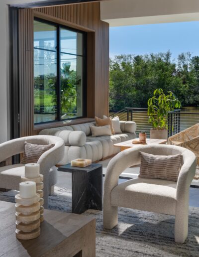 Modern living room with curved armchairs, a cushioned sofa, and a coffee table with candles, opening to a balcony with greenery and river views.