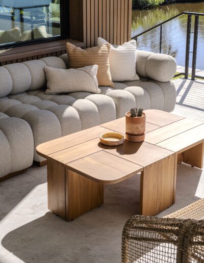 A modern outdoor seating area with a cushioned sofa, neutral throw pillows, a wooden coffee table with decor, and a woven chair beside a water feature.