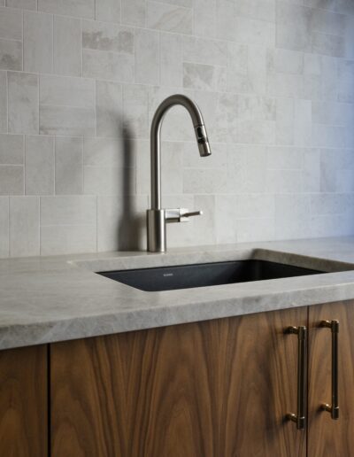 Modern kitchen sink with a brushed metal faucet, gray stone countertop, wood cabinets, and light gray tiled backsplash.