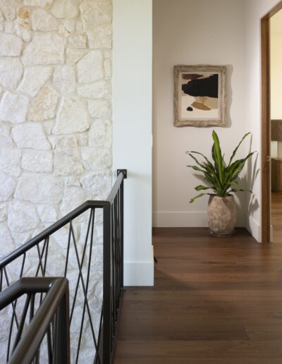 A hallway with a stone wall, black metal railing, wooden floor, potted plant, framed abstract art, and view into an adjacent room.