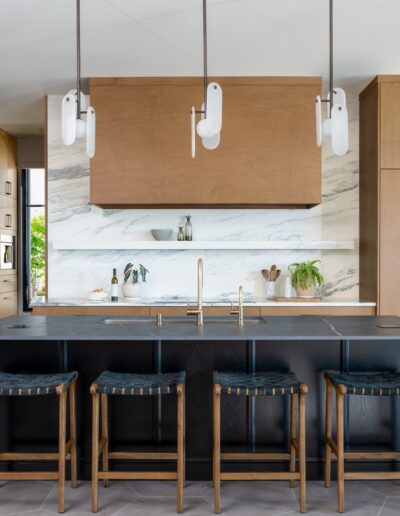Modern kitchen with wood cabinets, marble backsplash, black island with four bar stools, built-in ovens, and pendant lights hanging from the ceiling.