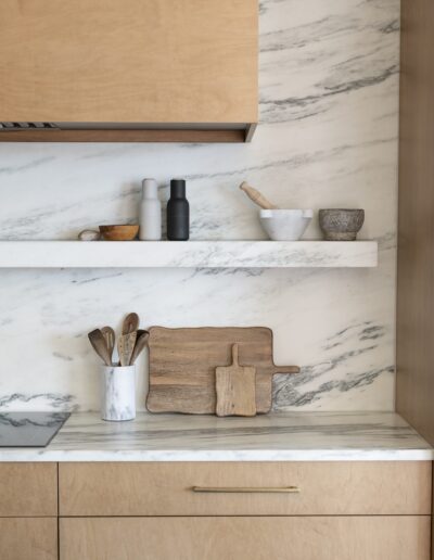 Minimalist kitchen with light wood cabinets, marble countertop and backsplash, a white shelf holding kitchenware, and wooden cutting boards arranged against the wall.